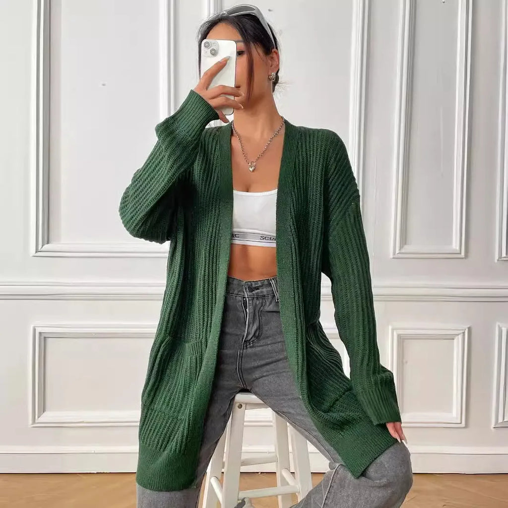 A woman wearing a loose-fitting, mid-length dark green cardigan sweater with double pockets.
