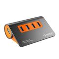 Dark grey aluminium USB hub with four ports, orange accents and detachable cable beside laptop