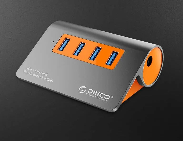 Dark grey aluminium USB hub with four ports, orange accents and detachable cable beside laptop