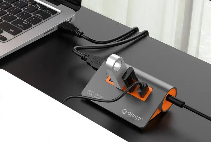 Dark grey aluminium USB hub with four ports, orange accents and detachable cable beside laptop