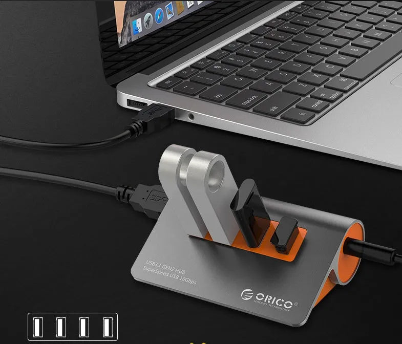 Dark grey aluminium USB hub with four ports, orange accents and detachable cable beside laptop