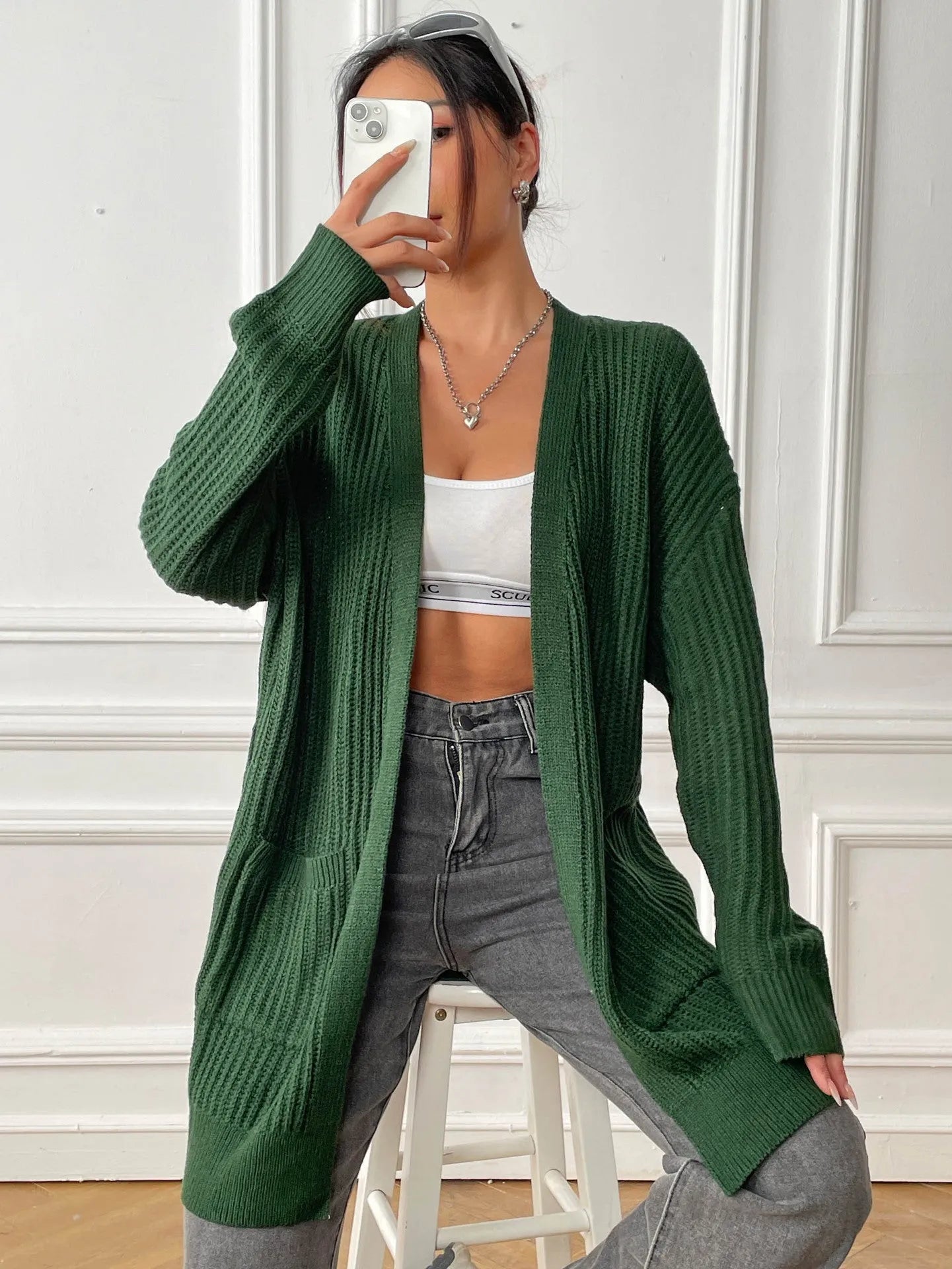 A woman wearing a loose-fitting, mid-length dark green cardigan sweater with double pockets.