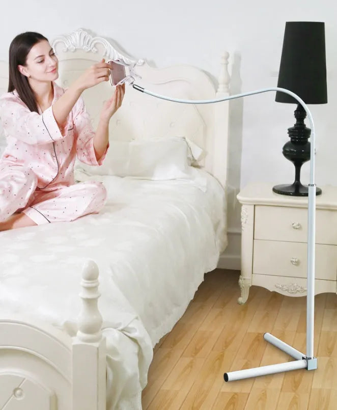 A black mobile phone bed bracket clipped to a bed frame, holding a smartphone in an overhead viewing position.