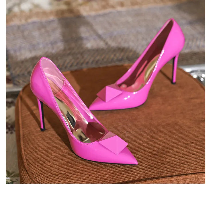 Pink women's pointed toe stiletto high heels with patent leather upper, rivet buckle detail, and a tall thin heel.