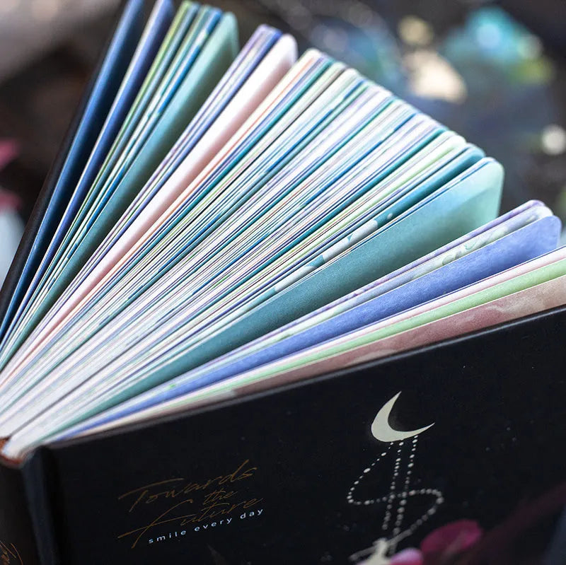 A5 notebook with a luminous cover open to show pages with colour illustrations, in deep blue, gold, light blue, or purple.