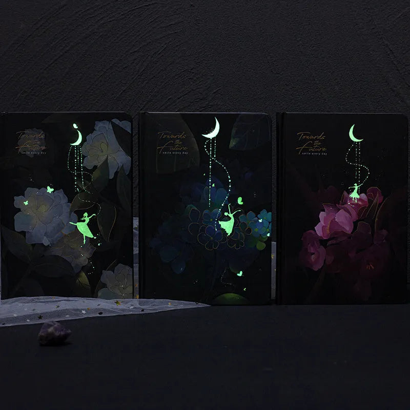 A5 notebook with a luminous cover open to show pages with colour illustrations, in deep blue, gold, light blue, or purple.