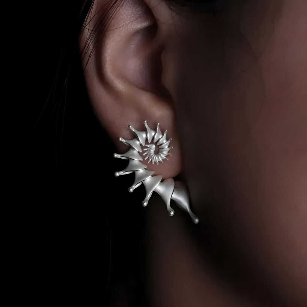 A pair of three-dimensional conch-shaped ear studs with a matte metallic finish.