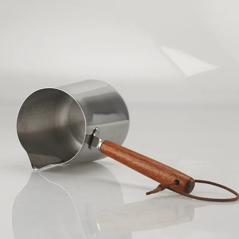 Stainless steel coffee mug with measurement scale and a light wood handle on a wooden table.