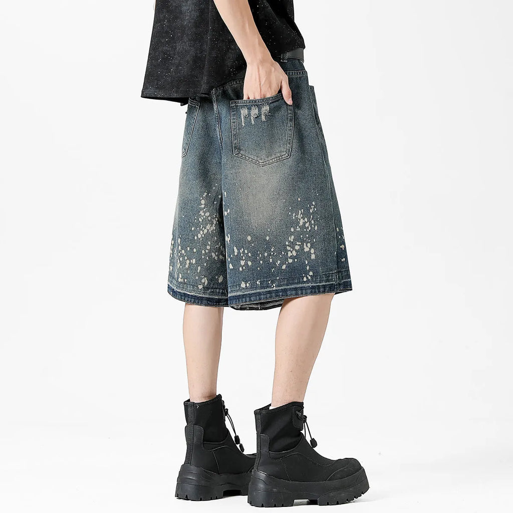Man wearing retro blue denim shorts with ripped and splash-ink distressing on a straight-leg cut.
