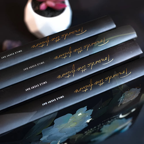 A5 notebook with a luminous cover open to show pages with colour illustrations, in deep blue, gold, light blue, or purple.